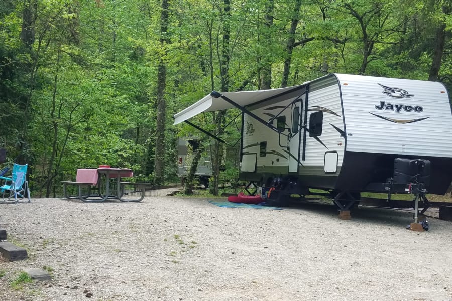 RV rental set up at a wooded campsite. A white Jayco travel trailer with an extended awning is parked on gravel, with a picnic area and chairs.