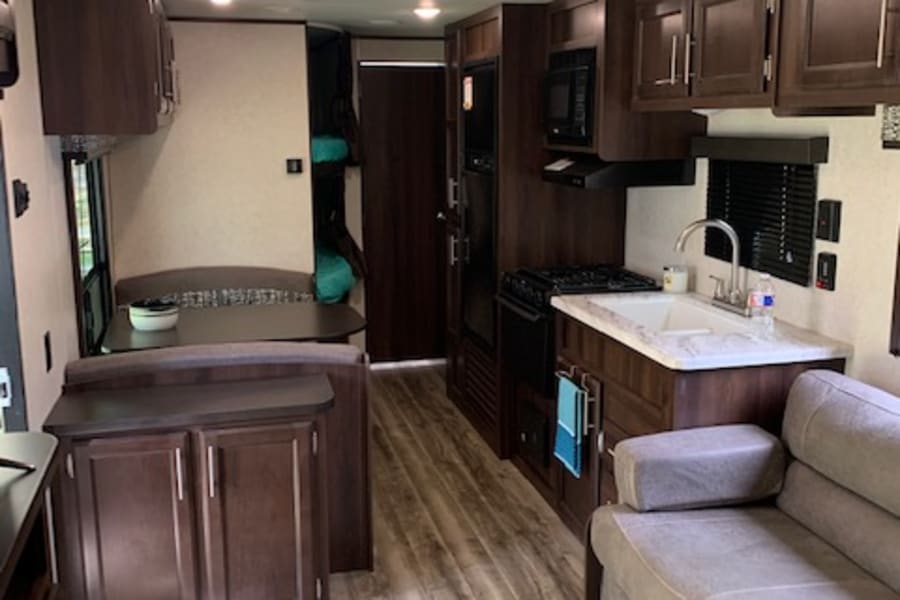 RV rental interior: modern kitchen with dark wood cabinets, light sofa, wood floor, and bed area. Brightly lit space.