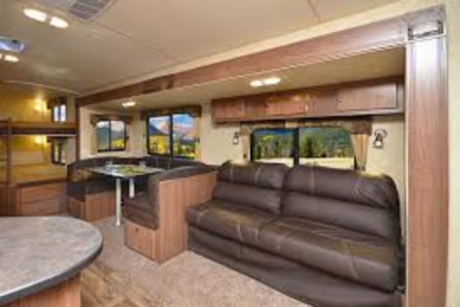 RV rental interior with brown sofa, dinette, and bunk beds. Large windows frame a scenic outdoor view. Ideal for comfortable travel.