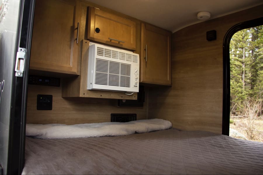 Inside a comfy RV rental: a bed, AC, wooden cabinets, & a window showing a peaceful forest view.