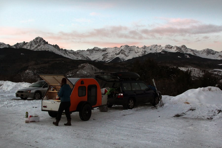 Scenic mountain RV rental trip: A person cooks at a teardrop trailer, parked with cars amidst snowy peaks.