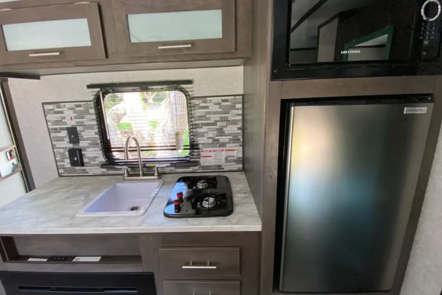RV rental's compact kitchen with stainless fridge, microwave, sink, 2-burner stovetop, and tiled backsplash.