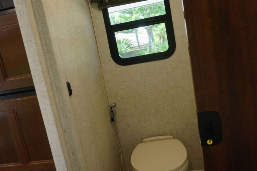 Compact RV rental bathroom: toilet, window with outdoor view, bidet sprayer, and overhead light.
