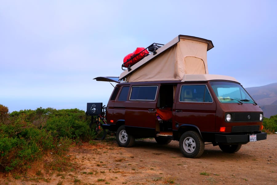 A brown pop-top camper van, loaded with gear and a bike, parked on a dirt road by the ocean. Perfect for an RV rental adventure.