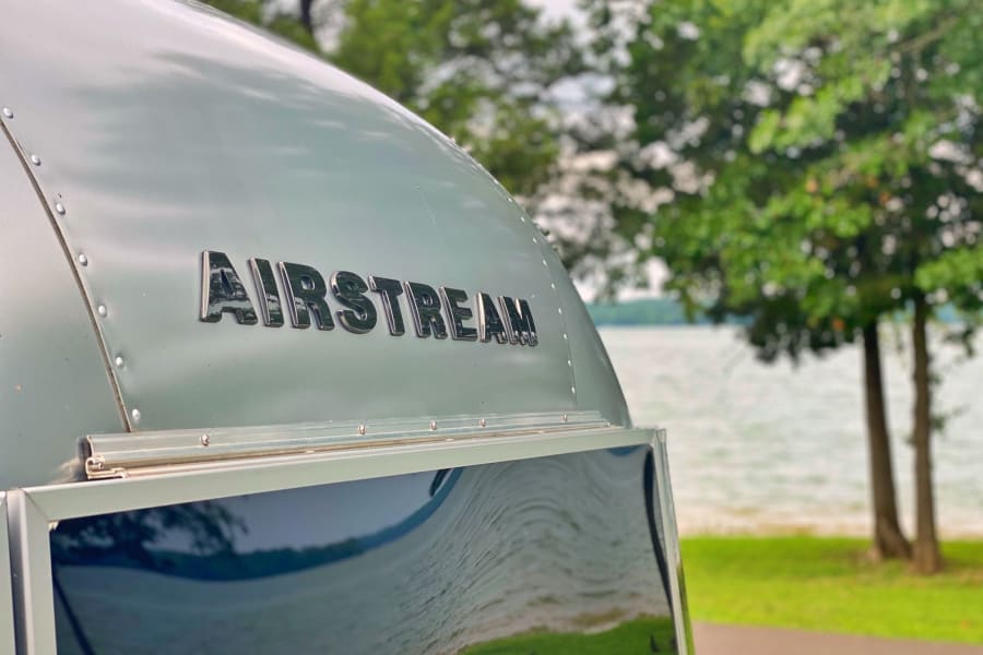 An Airstream RV, perfect for rv rental adventures, sits by a calm lake with trees in the background.