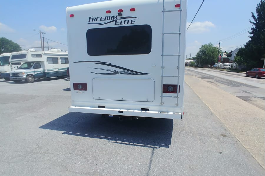 View the rear of a white Freedom Elite RV rental with a ladder & hitch, parked on an asphalt lot on a sunny day.