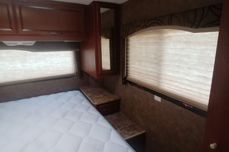 Cozy RV rental bedroom with a white mattress, overhead wood cabinets, and window blinds.