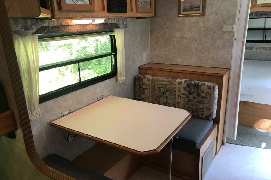 Cozy RV rental interior: A dinette with a table, booth seating, overhead cabinets, and a window overlooking lush greenery.