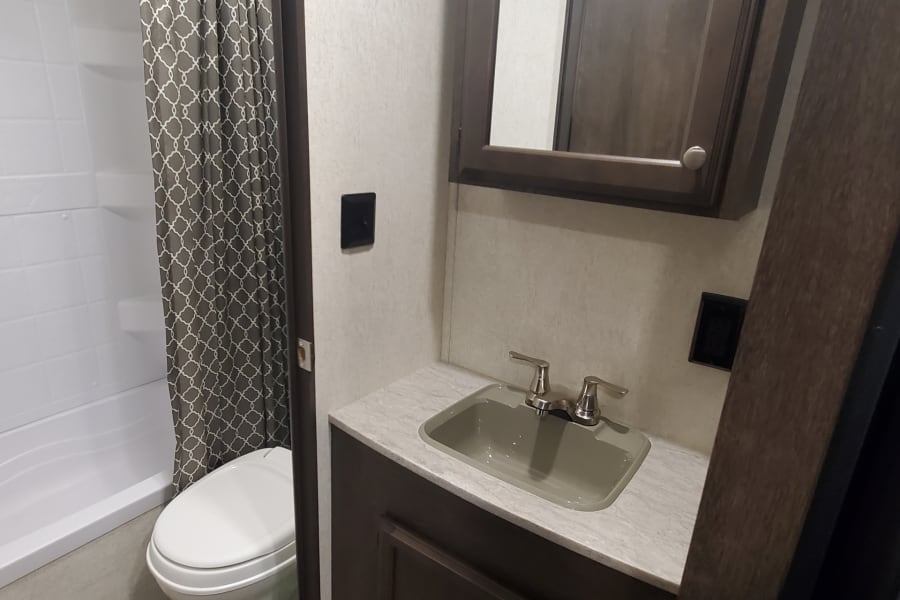 RV rental bathroom: white toilet, bathtub/shower with patterned curtain, vanity with sink and mirrored cabinet.