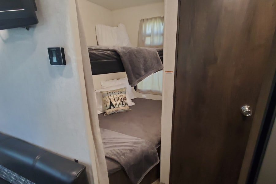 Inside an RV rental: a bunk bed area with grey blankets, a "Let's Go Camping" pillow, a black sofa, and a wall-mounted Dometic thermostat.