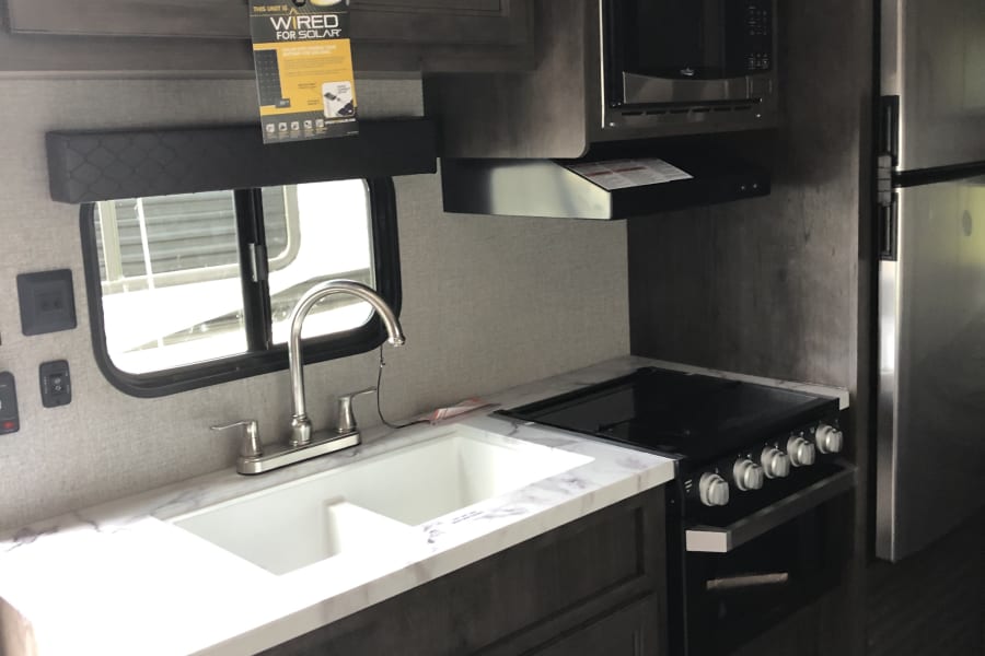 Modern RV rental kitchen with dark cabinets, white countertop, sink, stove, microwave, and fridge. Wired for solar.