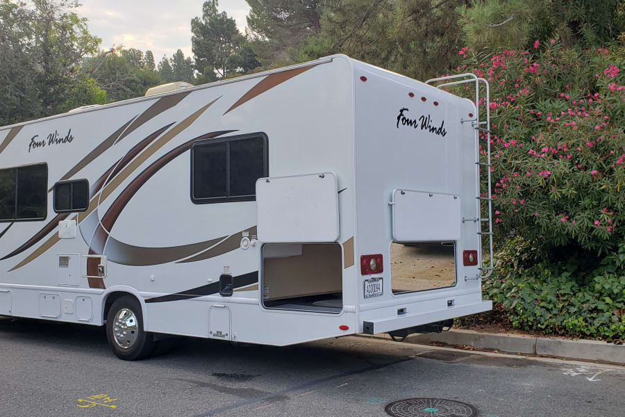 This white Four Winds RV rental, with brown graphics, is parked on a street near green bushes and pink flowers, storage open.