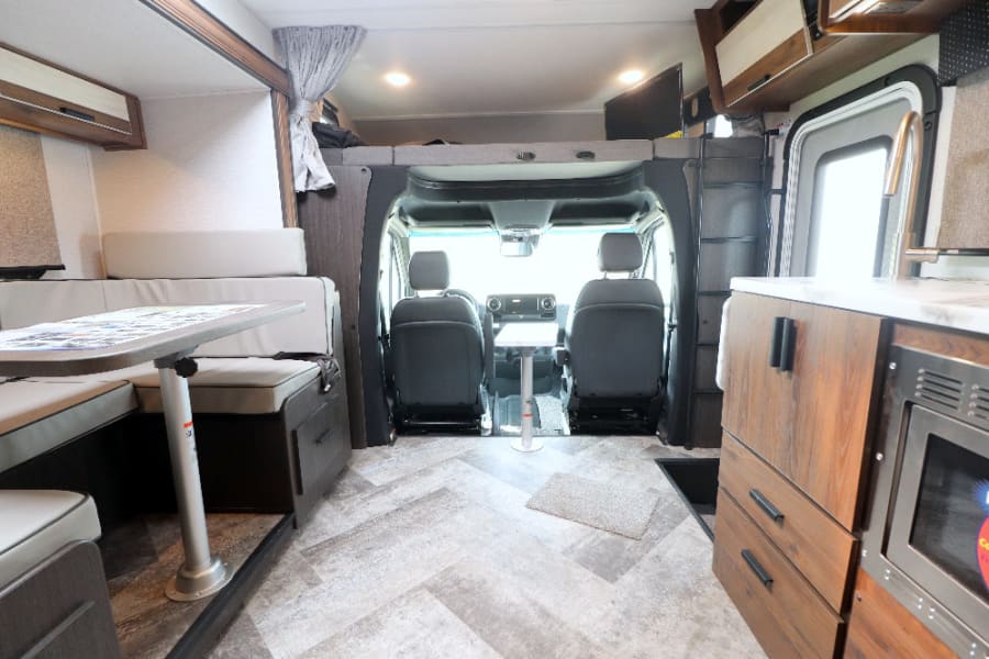 RV rental interior: driver's cabin, dining booth, kitchen counter with sink/microwave, and an overhead bed.