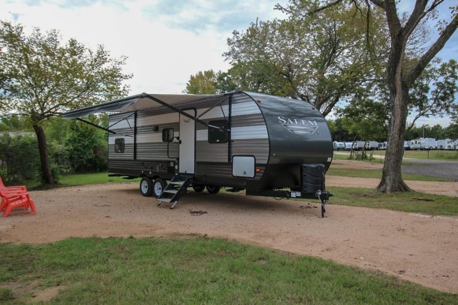 Experience camping with this spacious gray Salem travel trailer RV rental, parked on a gravel site under an extended awning, surrounded by trees.