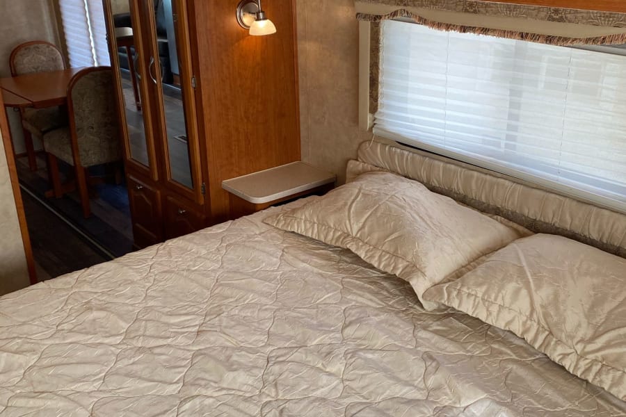 Inside an RV rental: a bright bedroom with a queen bed, wooden cabinets, and a mirrored closet. Dining area seen beyond.