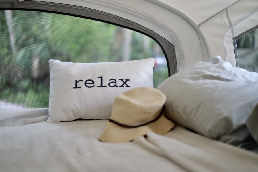 Relax in comfort! This cozy RV rental bed features a "relax" pillow and hat, with nature visible through the screen.