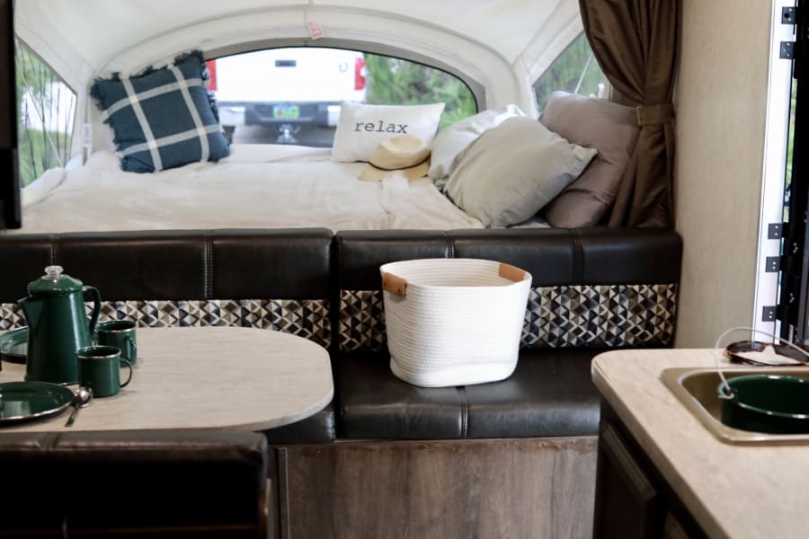 Cozy rv rental: comfy bed with 'relax' pillow, dining nook with greenware, plus a useful white storage basket.