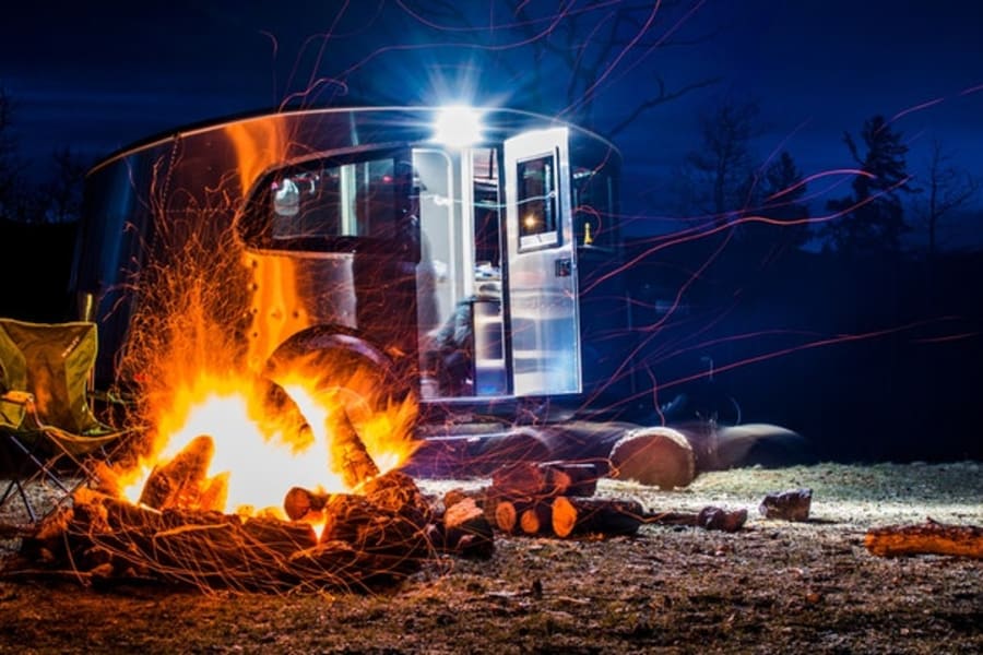 Cozy nighttime rv rental camping scene: a bright campfire with sparks, a chair, and a shining Airstream under dark skies.