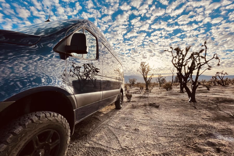 Dark adventure van reflects a vibrant cloudy sky, parked in a desert with sparse trees. Perfect RV rental escape!
