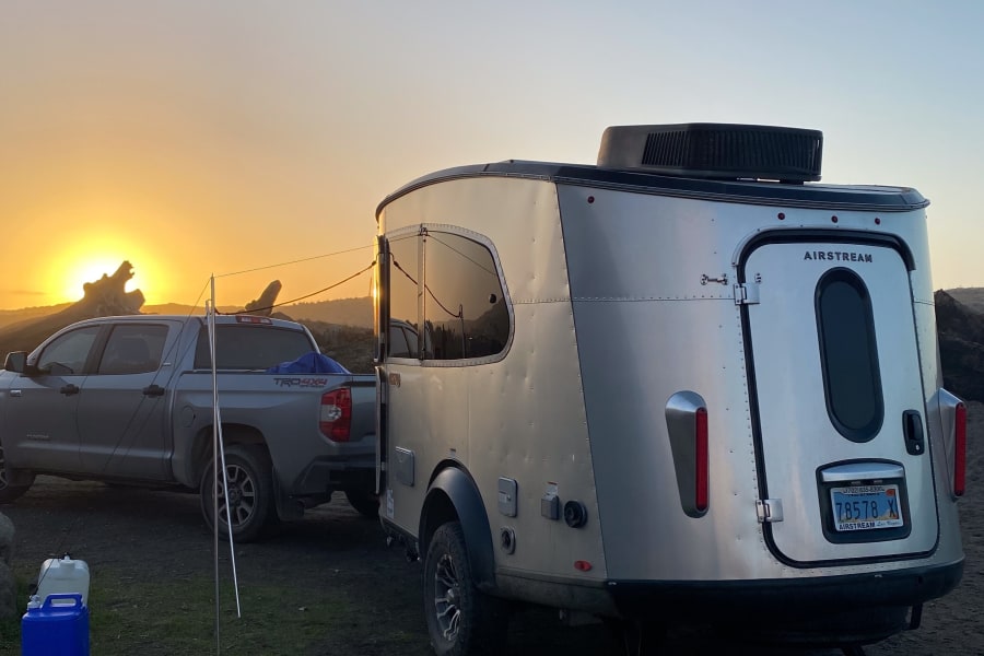 A silver Airstream travel trailer is hitched to a pickup truck at sunset, suggesting an ideal RV rental. Camping gear visible.