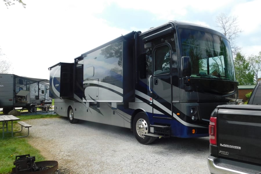 A large motorhome RV rental, with slide-outs extended, is parked on a gravel site near a travel trailer and pickup.
