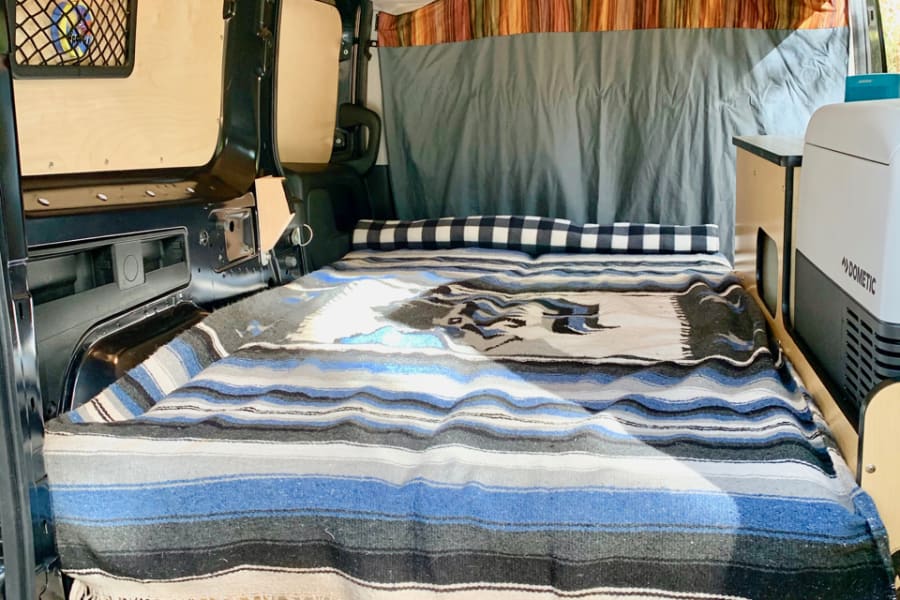 This RV rental's cozy camper van interior features a bed with storage, wood paneling, and a Dometic cooler.
