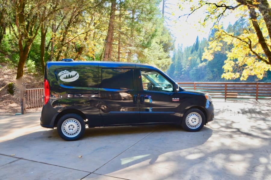 A black compact van, an RV rental, parked outdoors by a river, surrounded by vibrant forest. Ideal for travel adventures.