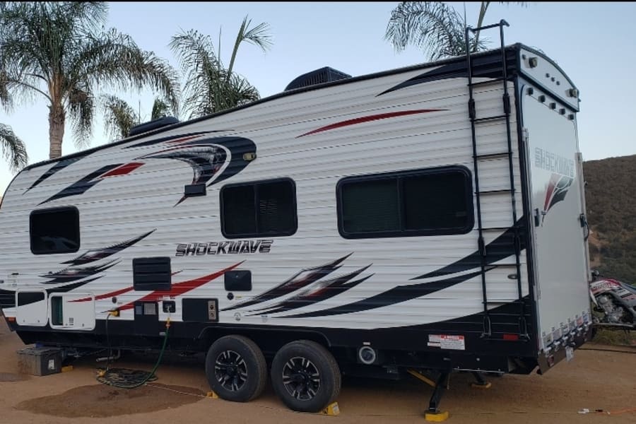 White Shockwave toy hauler RV rental, detailed with red/black graphics and a rear ladder, set up for outdoor adventure.