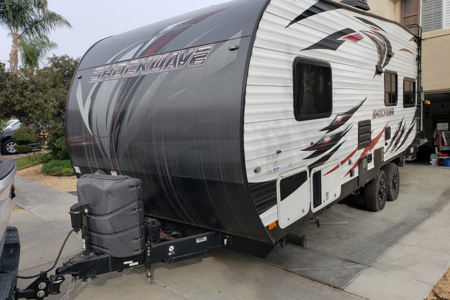 Large white and gray Shockwave travel trailer, ready for RV rental, parked on a paved driveway with hitch visible.