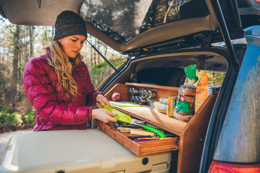 Woman prepares food using a custom portable kitchen in an SUV trunk, ideal for RV rental road trips.