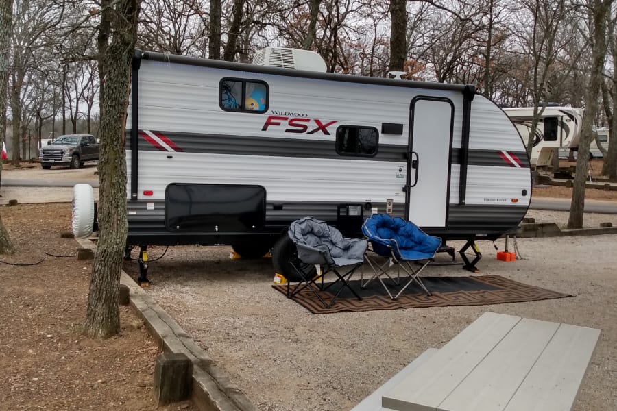 An RV rental travel trailer at a serene wooded campsite, featuring outdoor chairs and a picnic table.