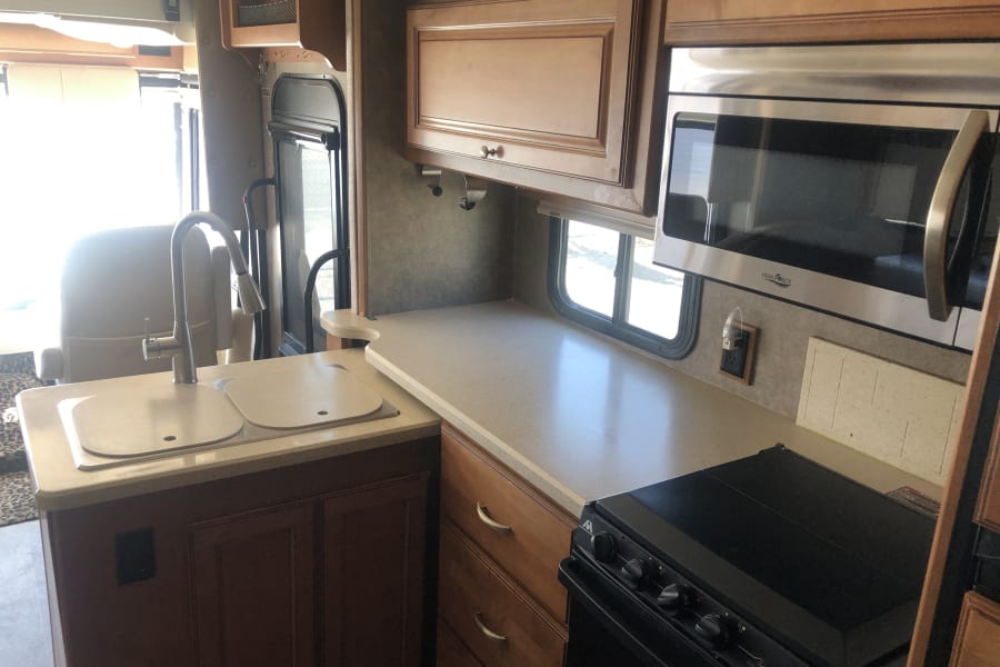 Bright RV rental kitchen with light countertops, dual sink, stainless microwave, black stove, and wood cabinets.
