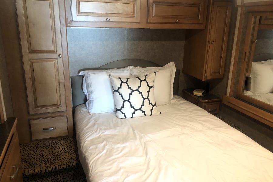 Inside a cozy RV rental: a comfortable bed with white linens, surrounded by wood cabinets & storage.