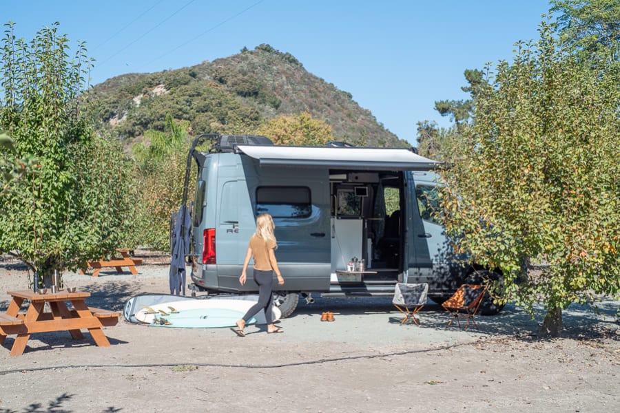Grey RV rental camper van parked with open side. A woman walks towards it. Surfboard, chairs & picnic tables nearby.
