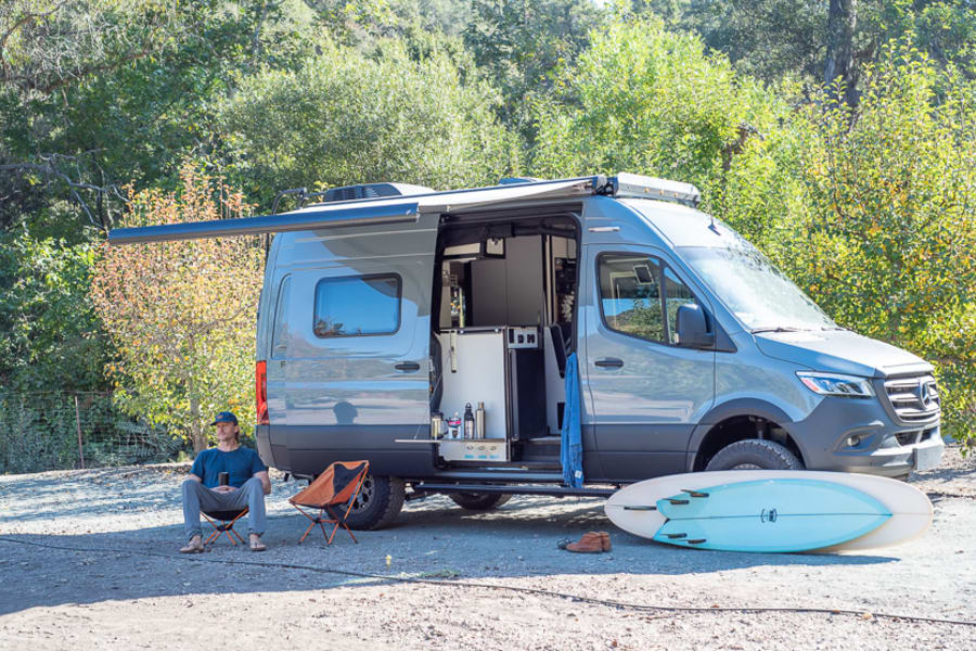 RV rental camper van, awning out, man relaxing on dirt next to surfboards. Ideal for outdoor adventures.