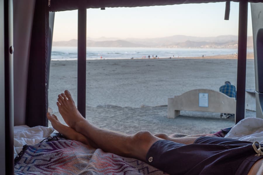 Enjoying an RV rental. Feet up, looking out the window at a sandy beach, ocean, and distant mountains. A serene view.