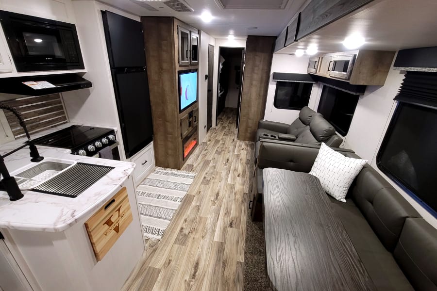 Modern RV rental interior featuring a compact kitchen, dinette, reclining sofa, TV, and fireplace in a clean, bright living space.