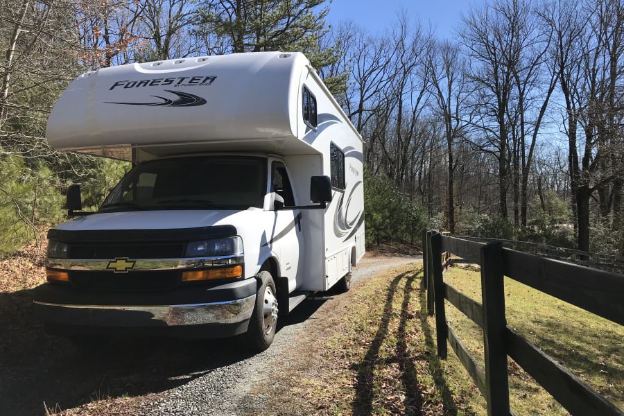 White RV motorhome on a gravel driveway, perfect for your next rv rental adventure. Surrounded by trees and a wooden fence.