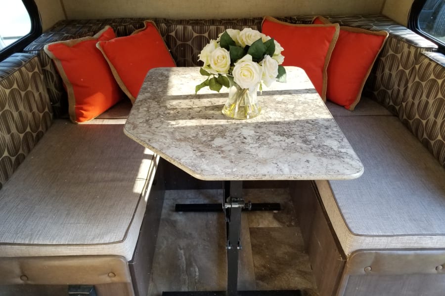 RV rental dinette: Cozy interior with gray bench seating, patterned backrests, orange accent pillows, and a table with flowers.