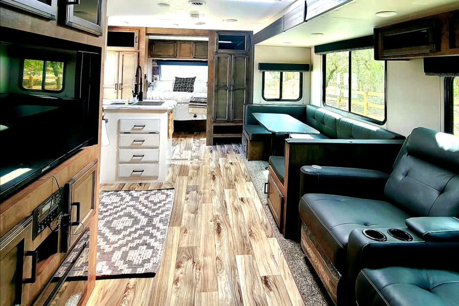 Cozy rv rental interior: dark recliners, dinette, and kitchen with wood-look flooring, leading to a comfortable bedroom.