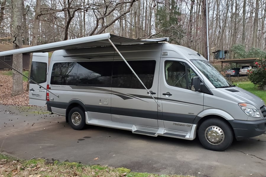 Silver Pleasure-Way RV rental with extended awning, parked on a driveway among trees. Perfect for adventure!