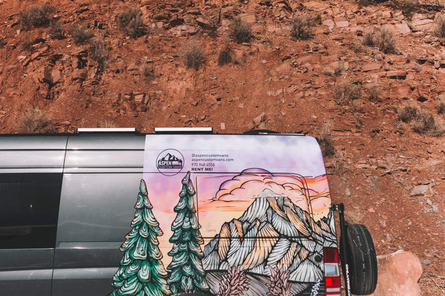 A grey custom rv rental van with a vibrant mountain mural, parked in snow by red rocks. Perfect for scenic adventures.