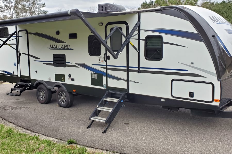 This white Mallard travel trailer, perfect for an RV rental, is parked with its awning extended and steps deployed.