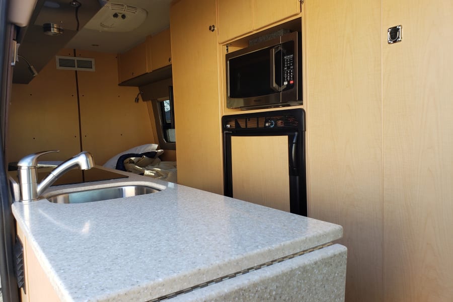 RV rental interior: kitchen with sink, light countertop, microwave, fridge, and a cozy bed area visible.
