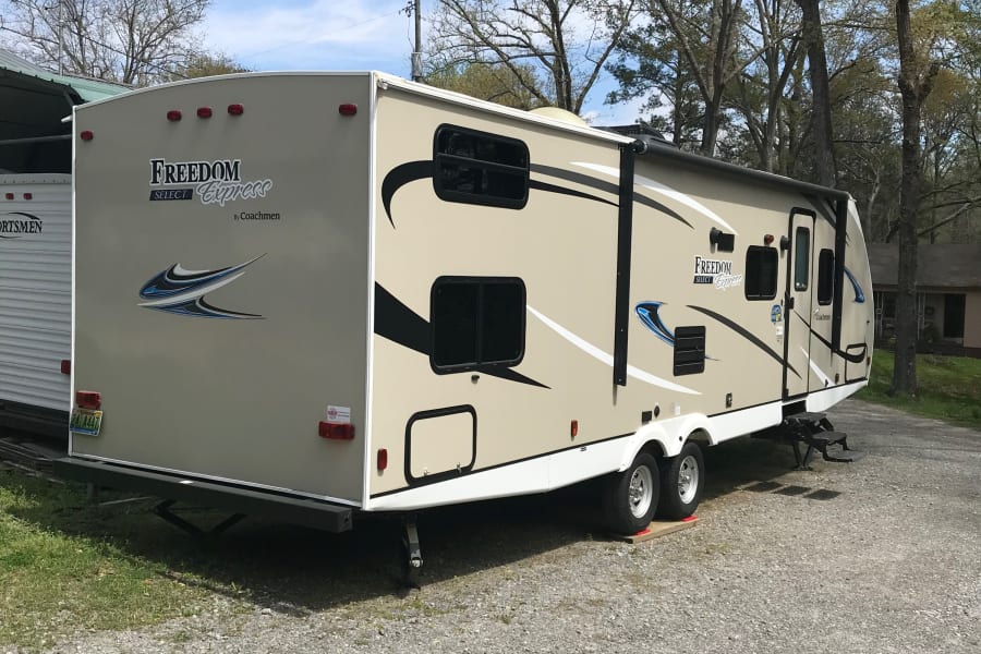 Tan Freedom Express travel trailer, an excellent RV rental option, parked on gravel under a blue sky, surrounded by trees.