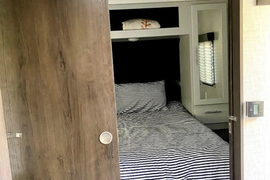 Inside an RV rental: a compact bedroom with a striped bed, built-in storage, and a partial view through the sliding door.