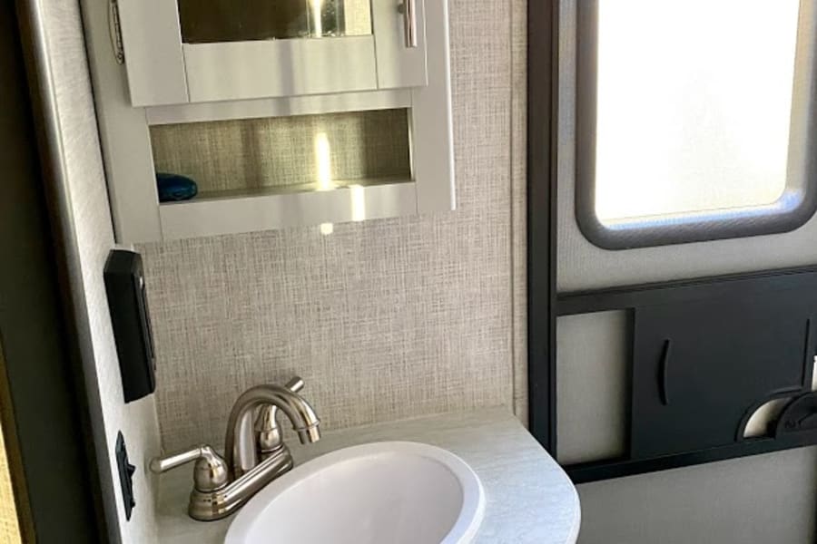 RV rental bathroom features a compact sink, faucet, white vanity, wall cabinet, and brightly lit door.