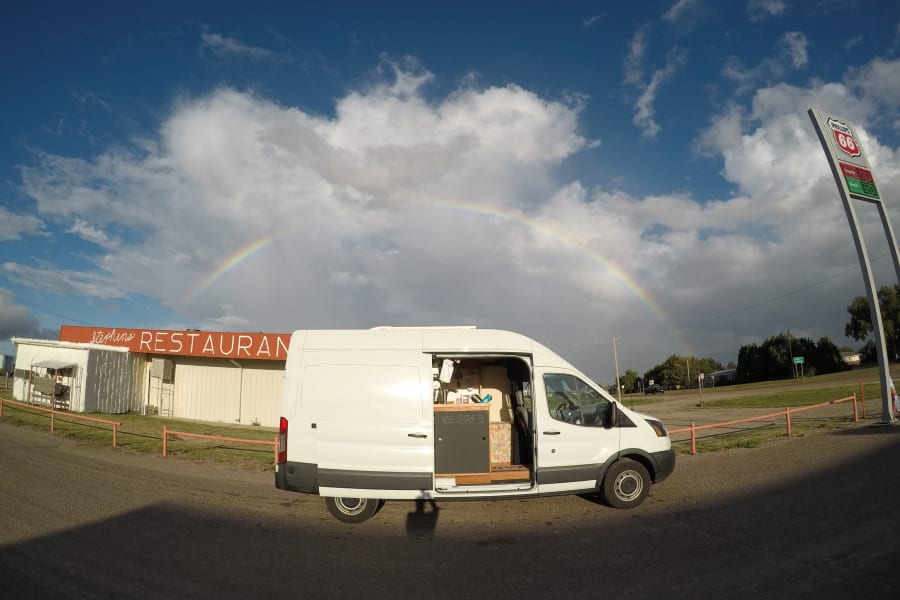 White camper van (RV rental) under a vibrant rainbow near Stephens Restaurant and Phillips 66 sign.