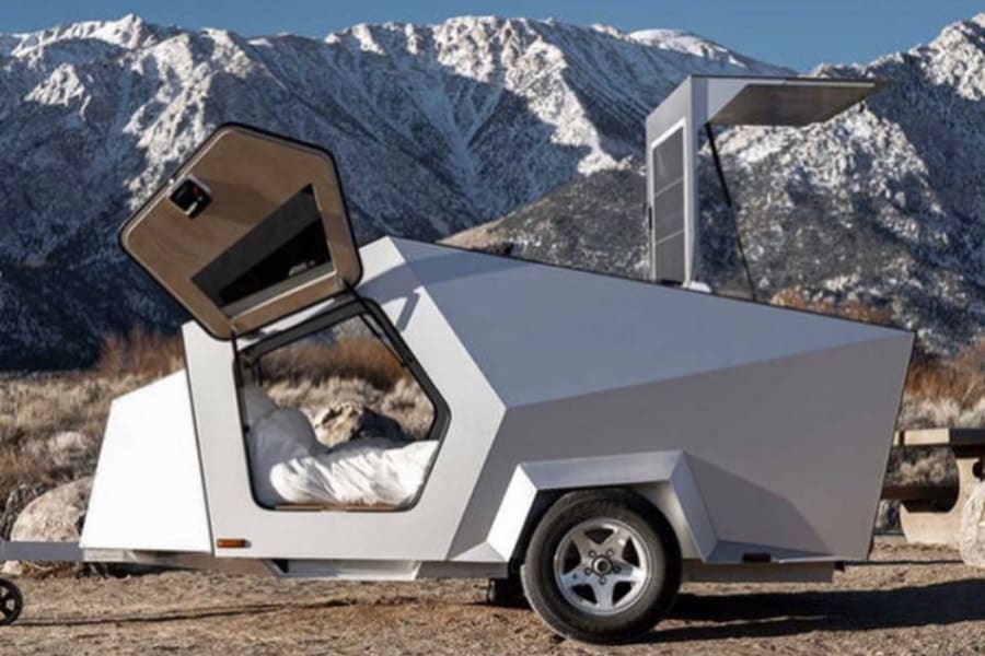 A futuristic, angular travel trailer, perfect for an RV rental, is parked among snow-capped mountains. A bed is visible inside.