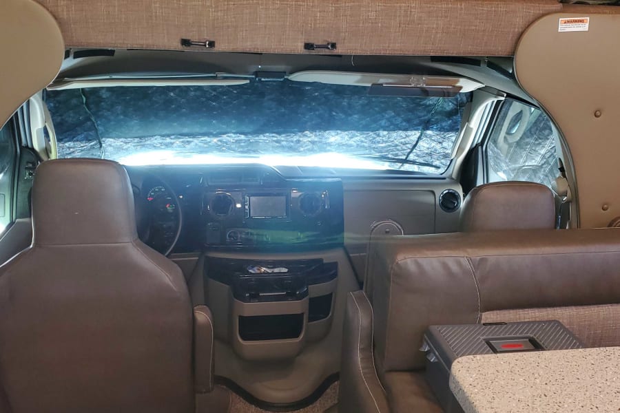 RV rental interior: The front cab area with dashboard, steering wheel, seats, and covered windshield, from the living space.